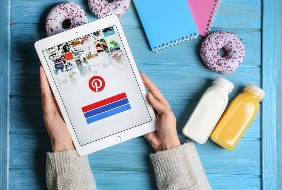 Hands holding tablet with Pinterest icon on blue background with purple donuts and drinks