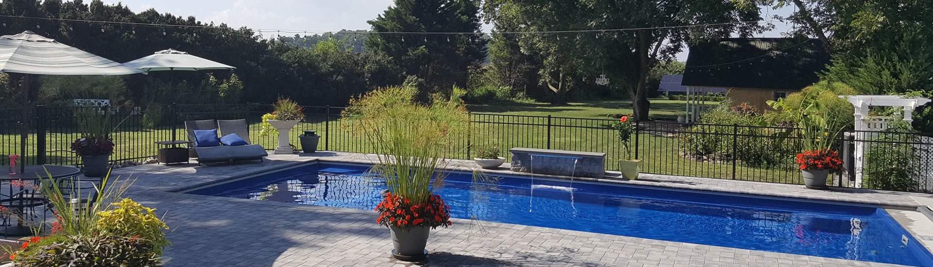 Blue swimming pool sourrounded by an iron fence with tall trees and grass lawn in the background.