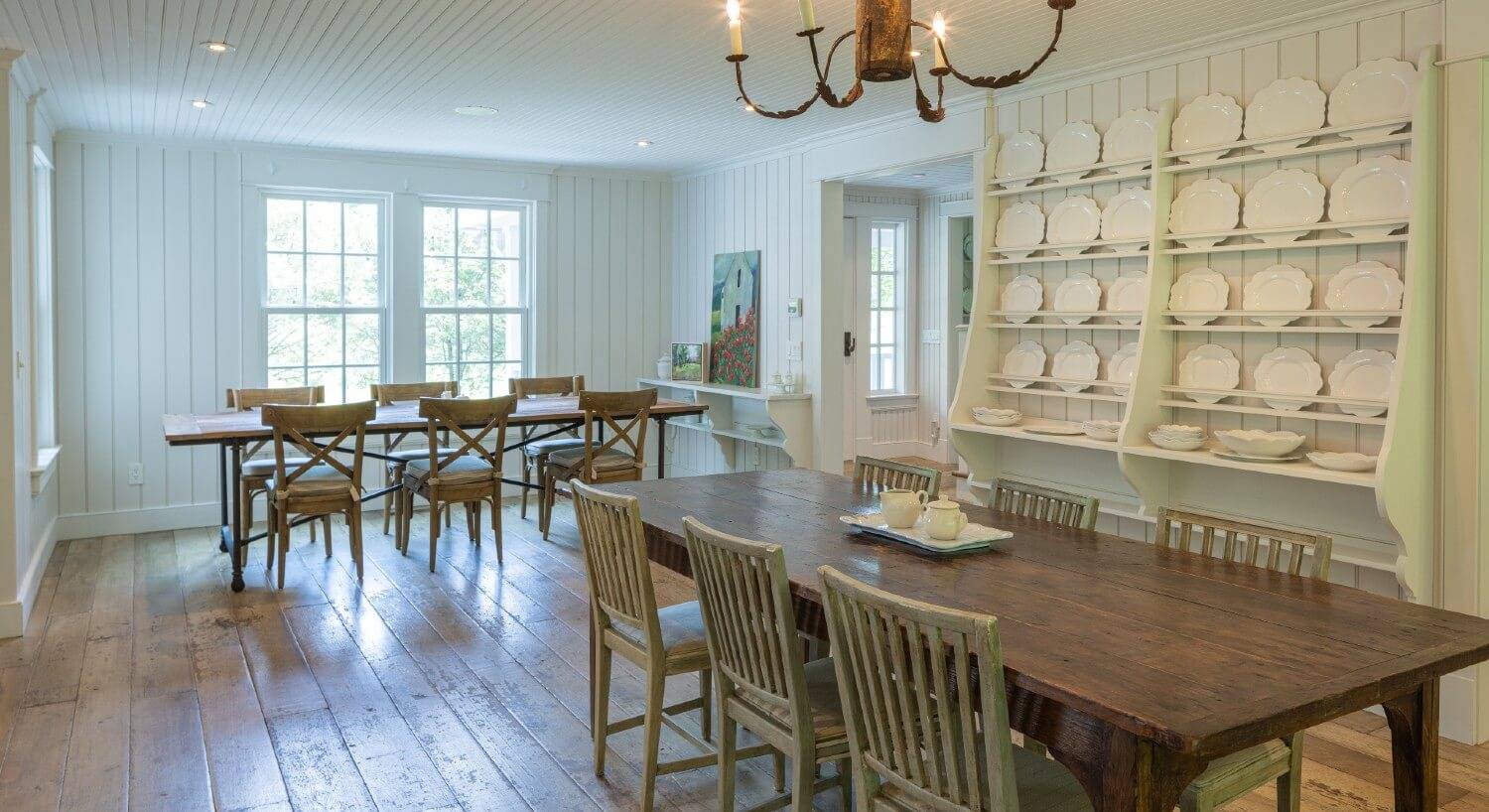 Large dining room of a home with two tables for six and plate rack on a wall full of white plates