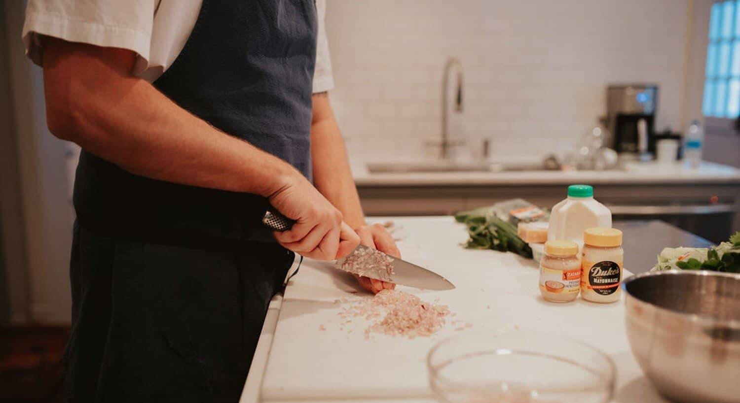 A chef in a blue apron dicing onion on a white cutting board with bowls and condiments on the counter
