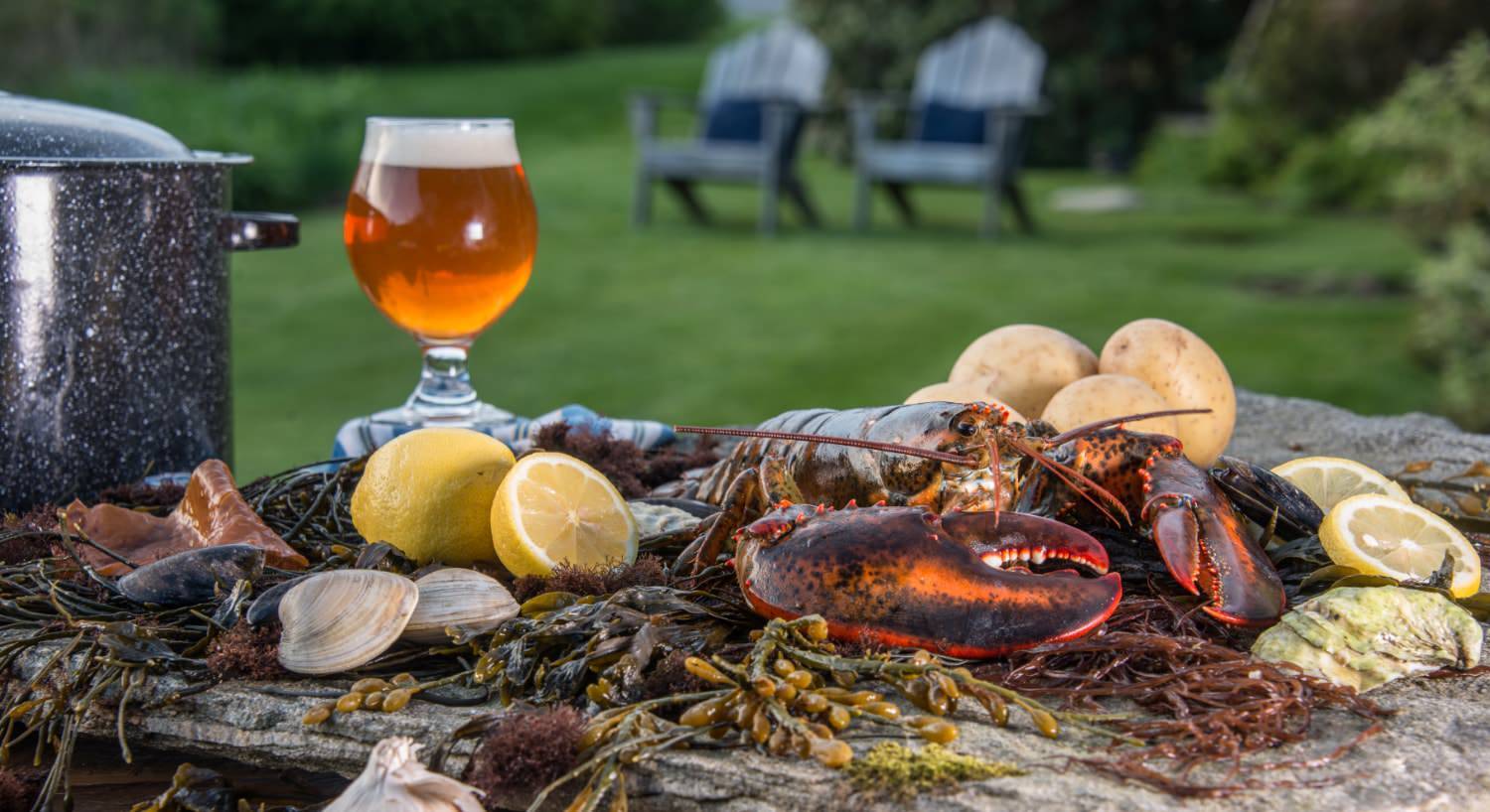 Close up view of large flat rock filled with lobster, clams, potatoes, and lemon slices and a glass of beer and large pot on the side