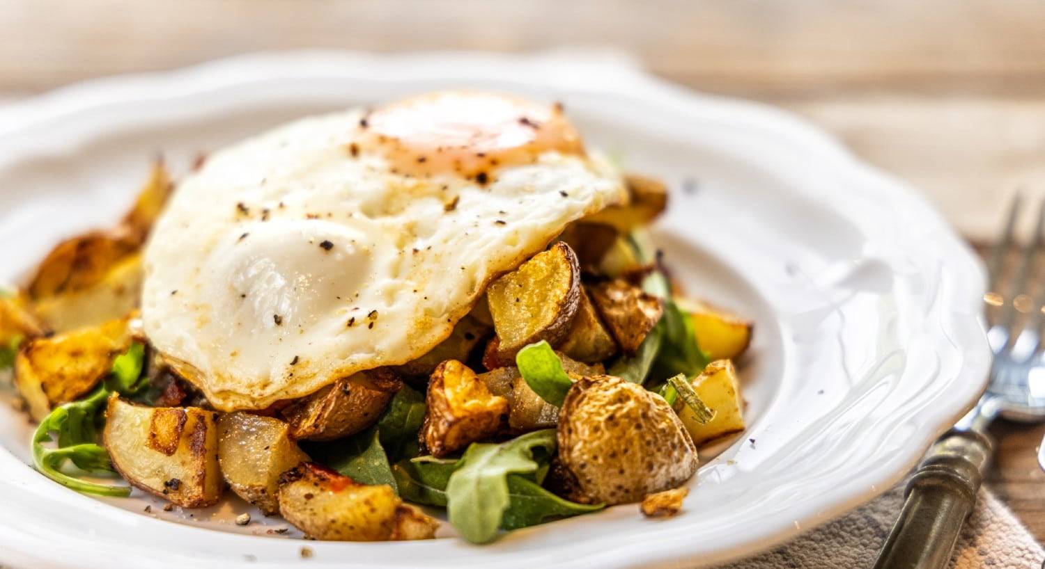 A breakfast hash of diced potatoes and greens with two fried eggs on a decorative white plate