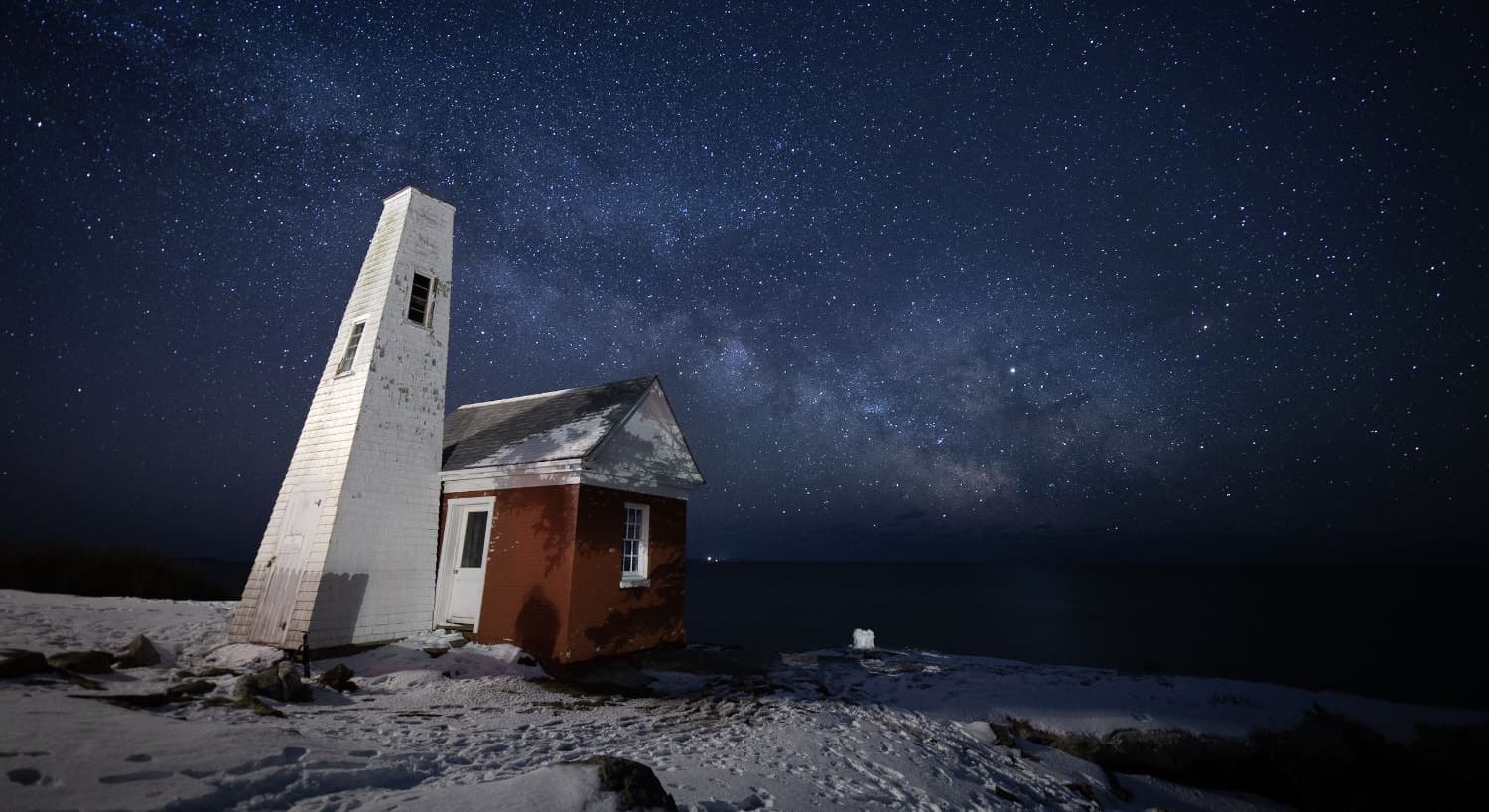 Small lighthouse surrounded by snow near the ocean at night with the sky full of stars
