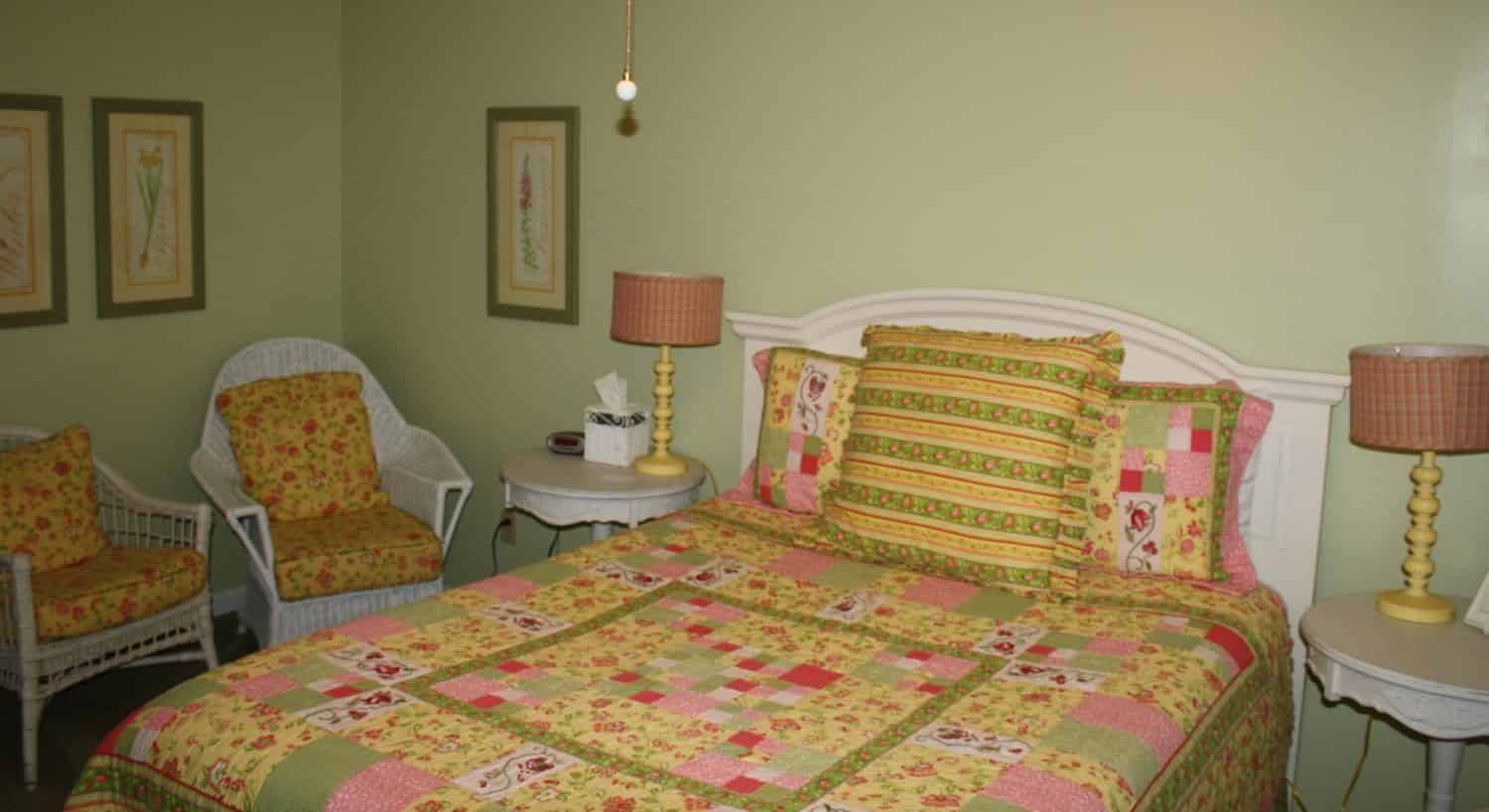 Bedroom painted light green with white headboard and green, yellow, and pink bedding and white wicker chairs