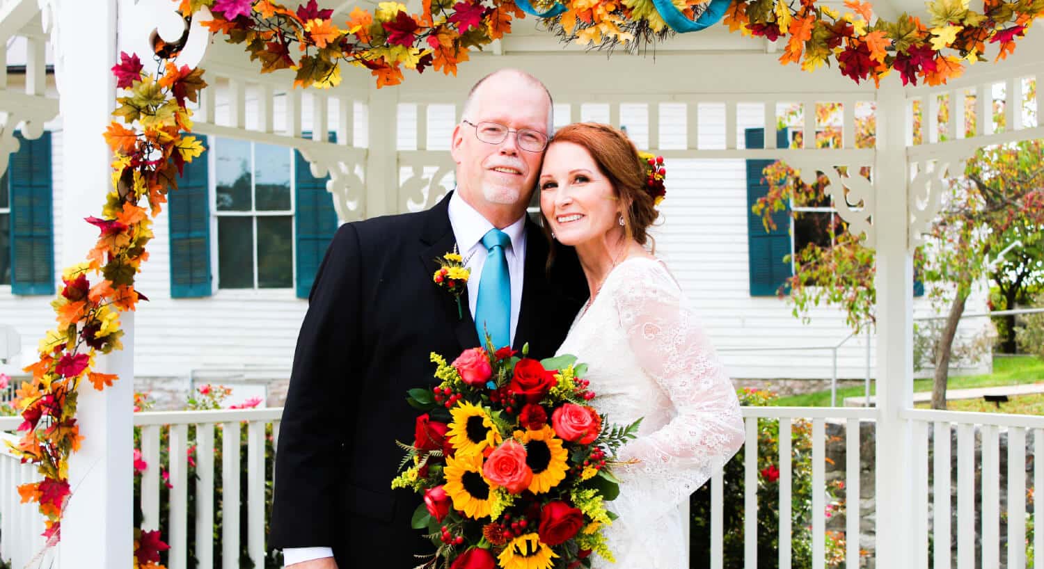 bride holding bouquet with fall flowers and groom standing by white gazebo  with draping fall flowers house with green shutters in distance
