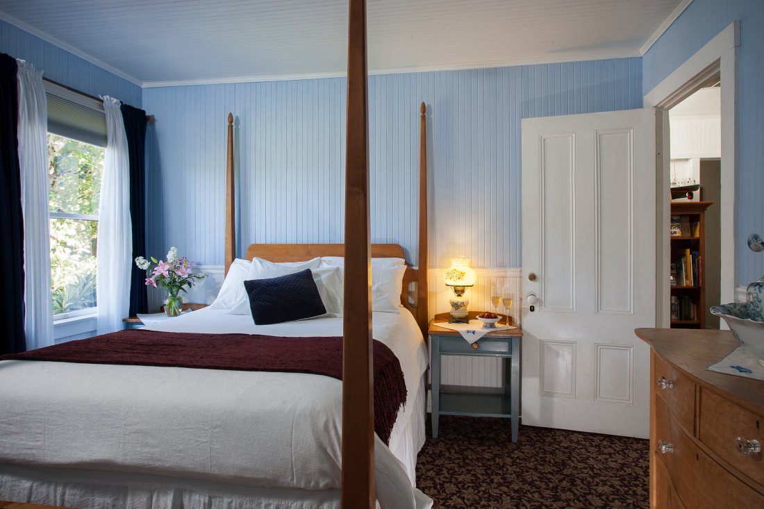 High post bed underneath white beadboard walls. Bedside tables flank bed draped in white, with flowers outside window. 