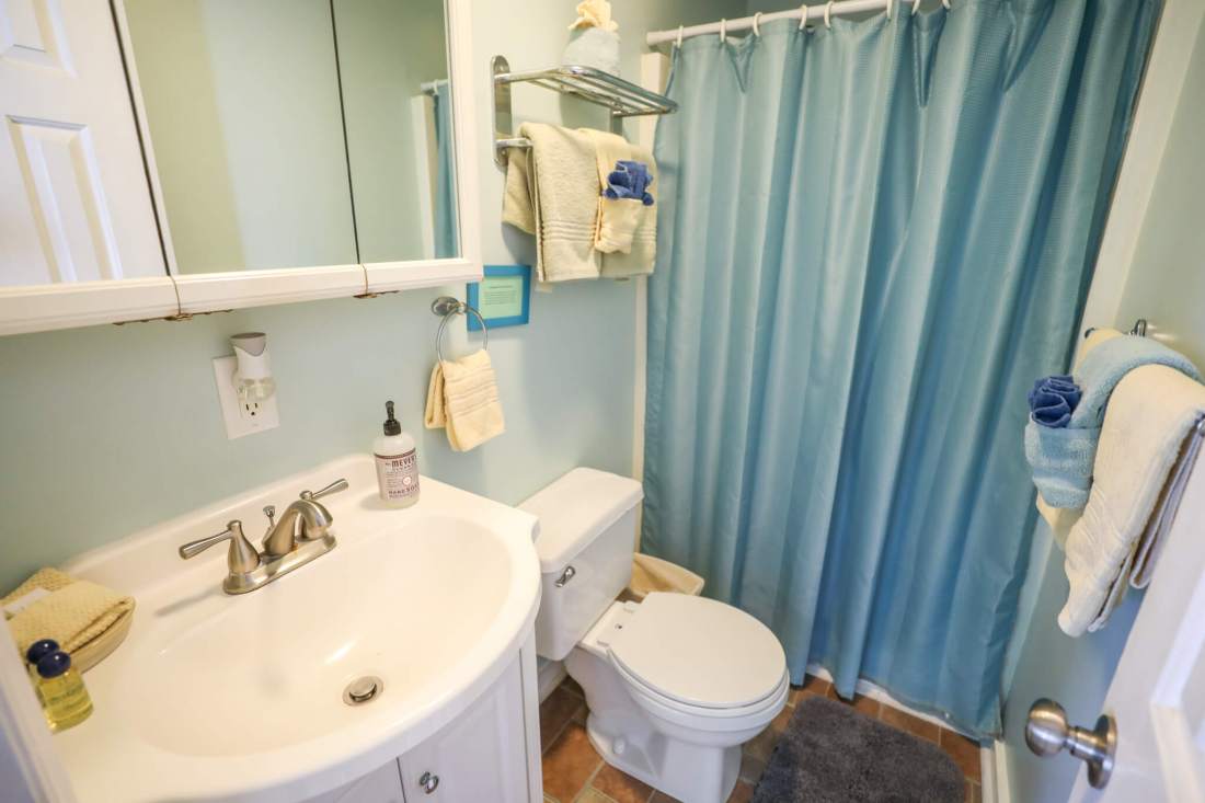 Interior view of bathroom with cabinet sink, blue shower curtain and beige and cream towels underneath wall mirror. 