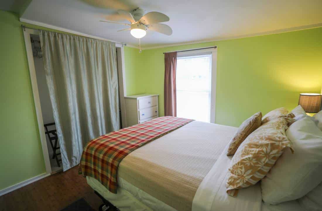 Interior view of bedroom decorated in greens, grays and reds. Bed underneath tall window in corner. 
