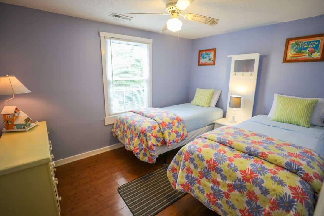 Interior view of bedroom with purple walls above twin beds underneath a window, and across from a dresser on wooden floors.