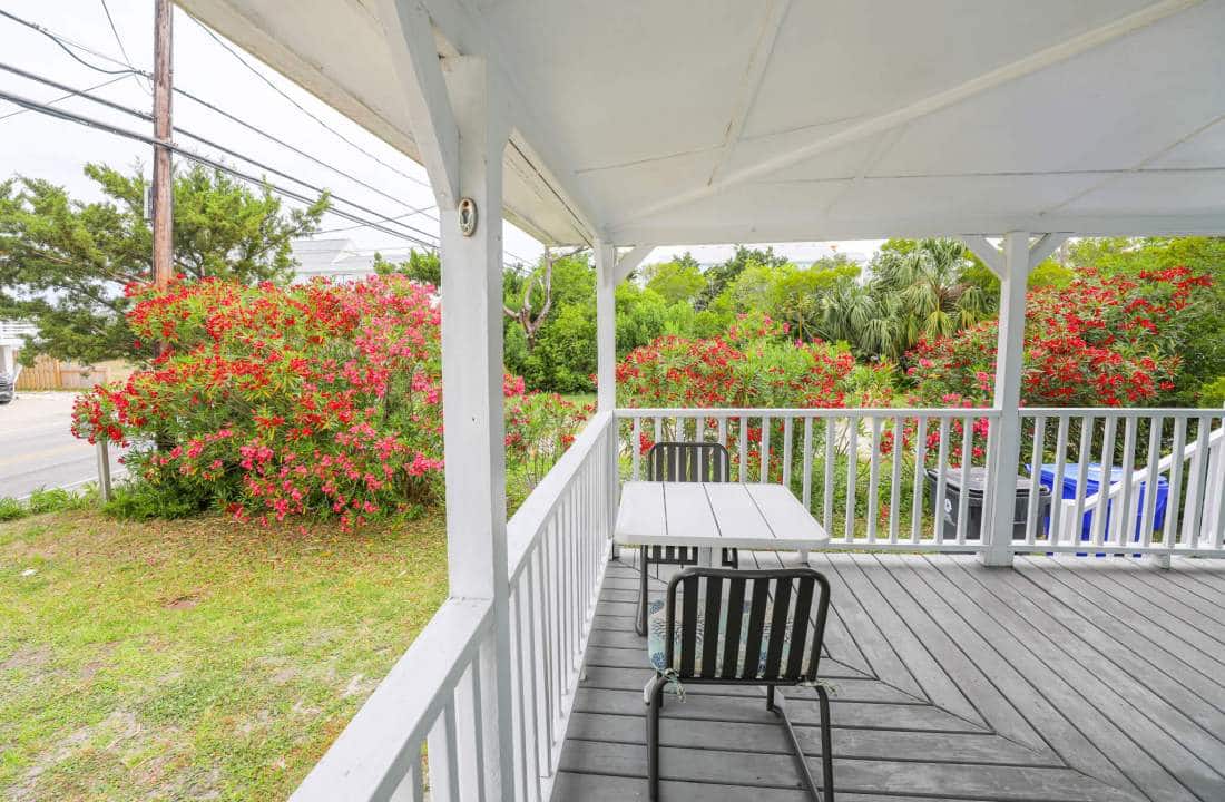 Exterior view of wraparound porch with patio chars around table. Flowering bushes in background. 