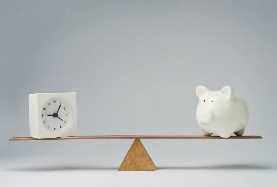 White alarm clock and white piggy bank on a see-saw, representing time vs. money